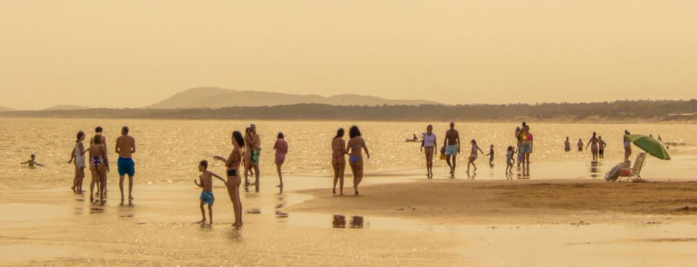 People enjoying a sunny day walking along a wide sandy beach with mountains visible in the distance. Landscape.