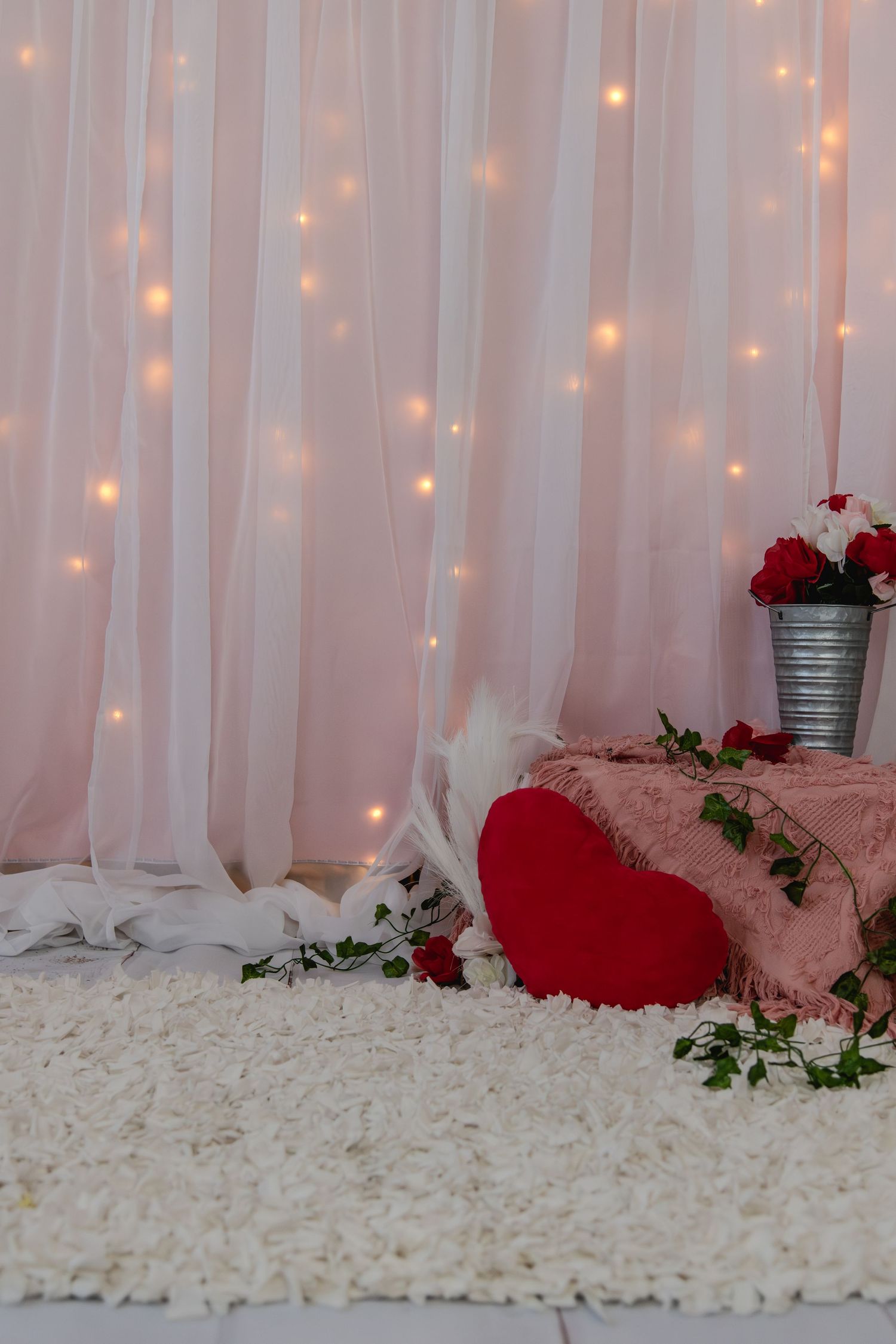 Romantic backdrop with sheer white curtains, twinkling string lights, red roses, and plush pillows on white shag carpet.