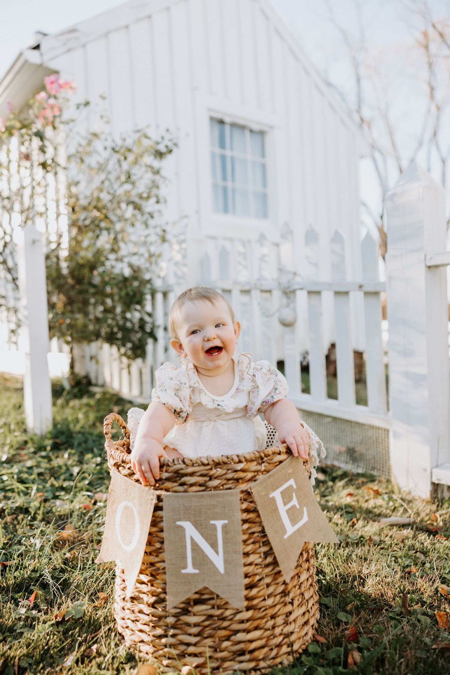 A small child sits in a woven basket outdoors with a burlap banner against a white picket fence backdrop.