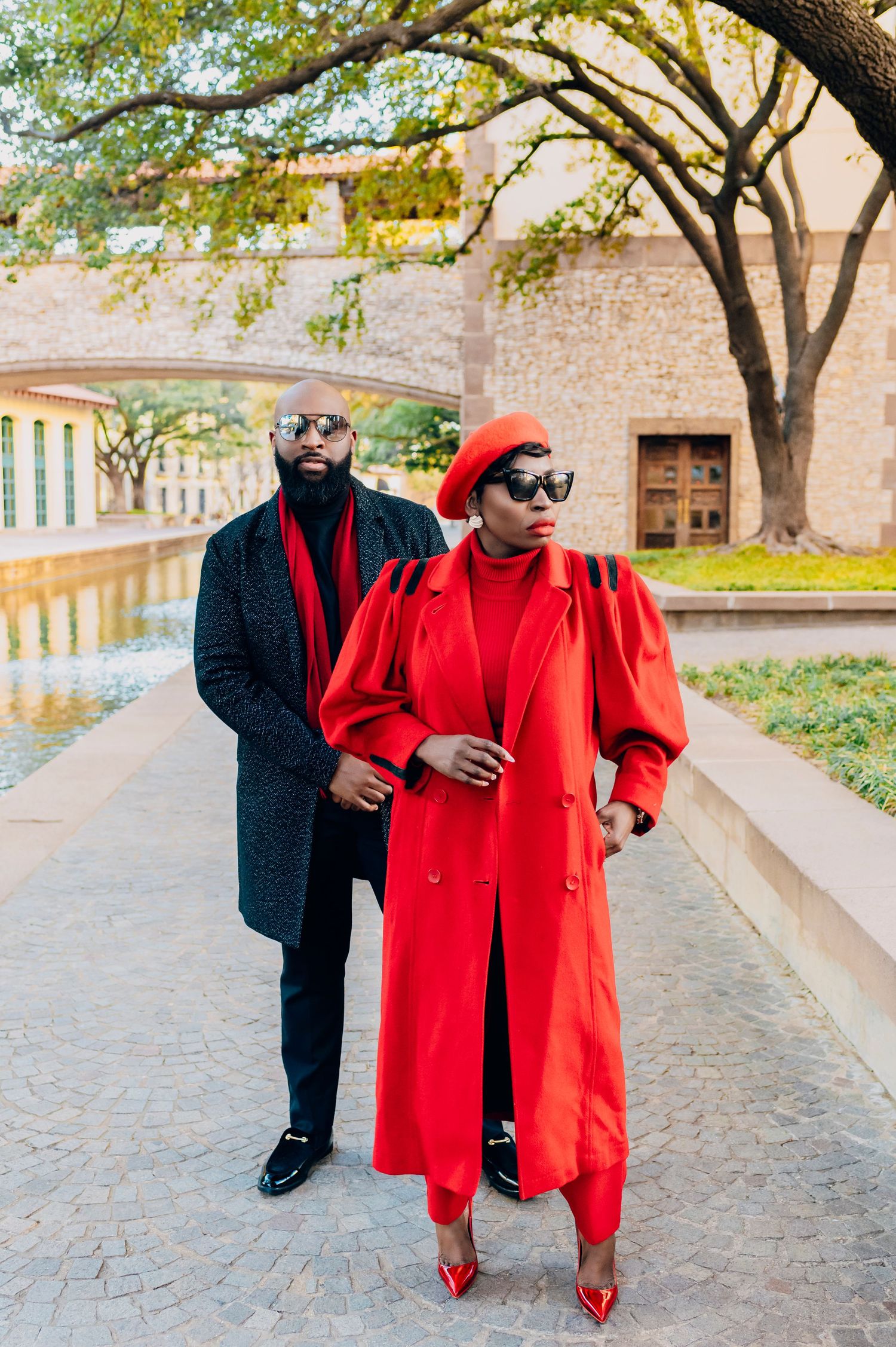 Two people in coordinated winter outfits pose outdoors, with a dramatic red coat and black ensemble against stone architecture.