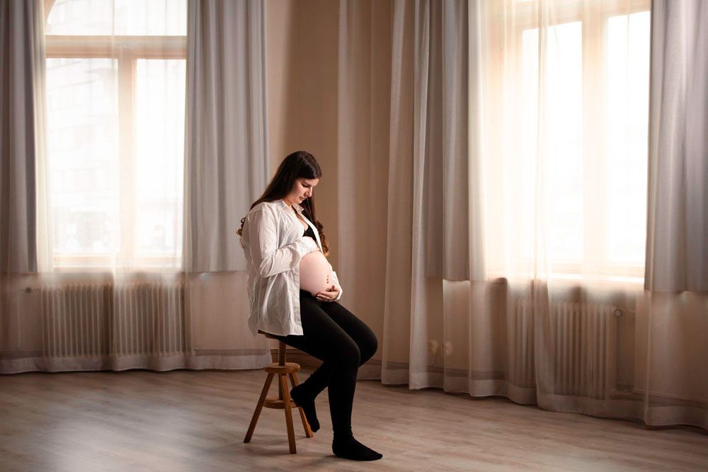 Valokuvausstudiolla otettu kuva, jossa raskaana oleva nainen istuu tuolilla, pitää hellästi raskausmahastaan kiinni ja katsoo sitä.