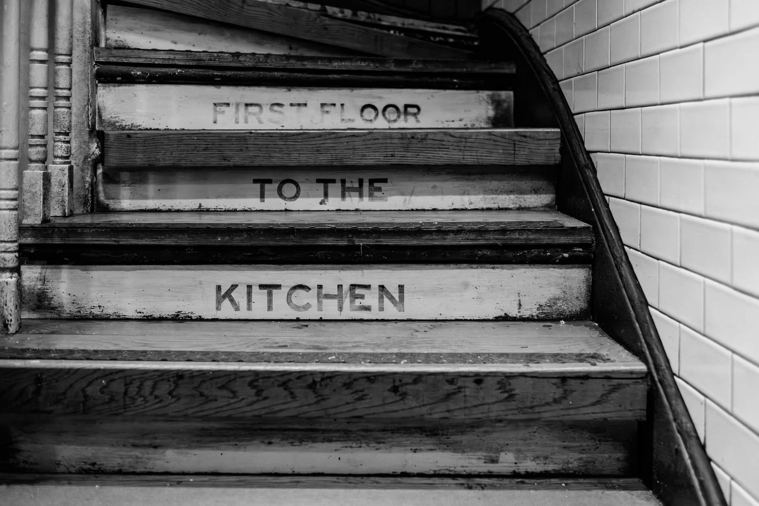 Black and white photograph of worn stairs with text reading 'First Floor' and 'To The Kitchen' painted on the steps.
