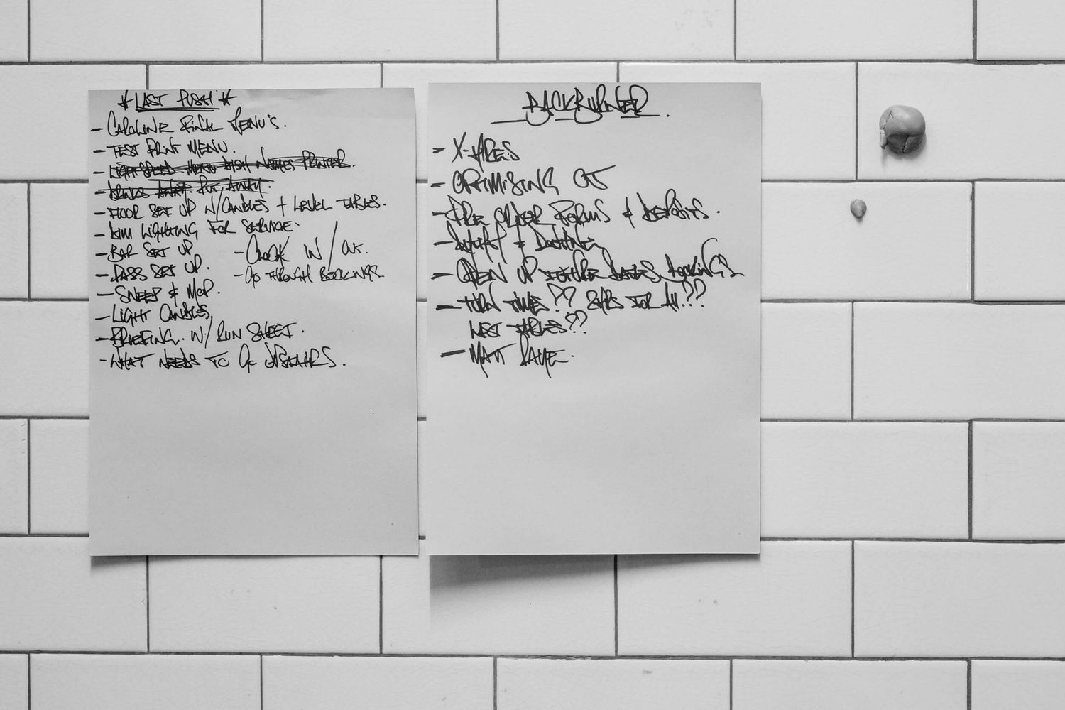 Handwritten lists on white paper posted against subway tile wall with small round fixture visible.