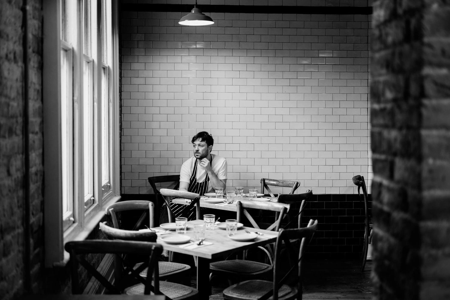 Black and white image of a minimalist restaurant interior with brick walls and window lighting.