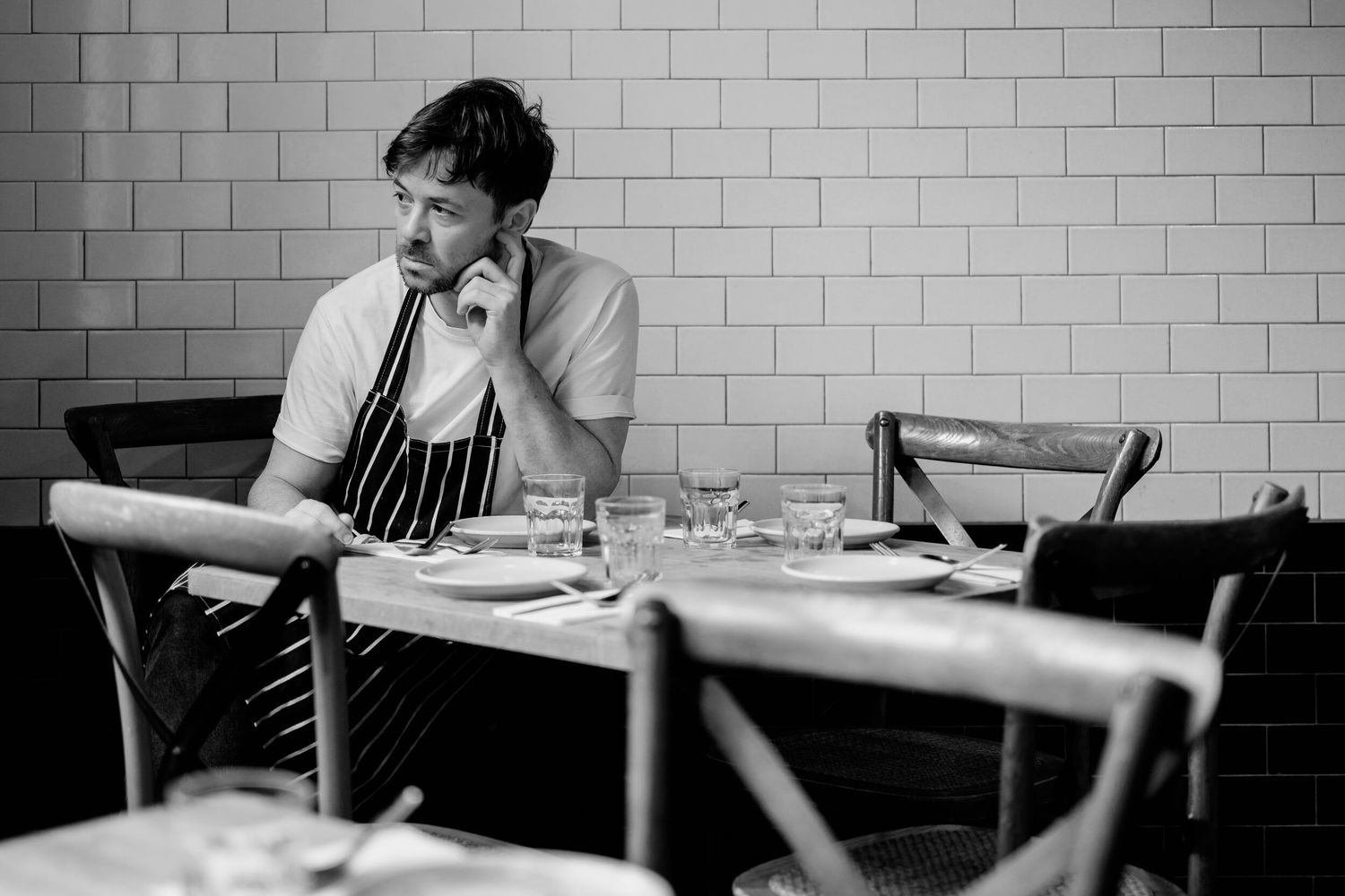A person sits alone at a table in a monochromatic restaurant setting with brick walls and natural light.