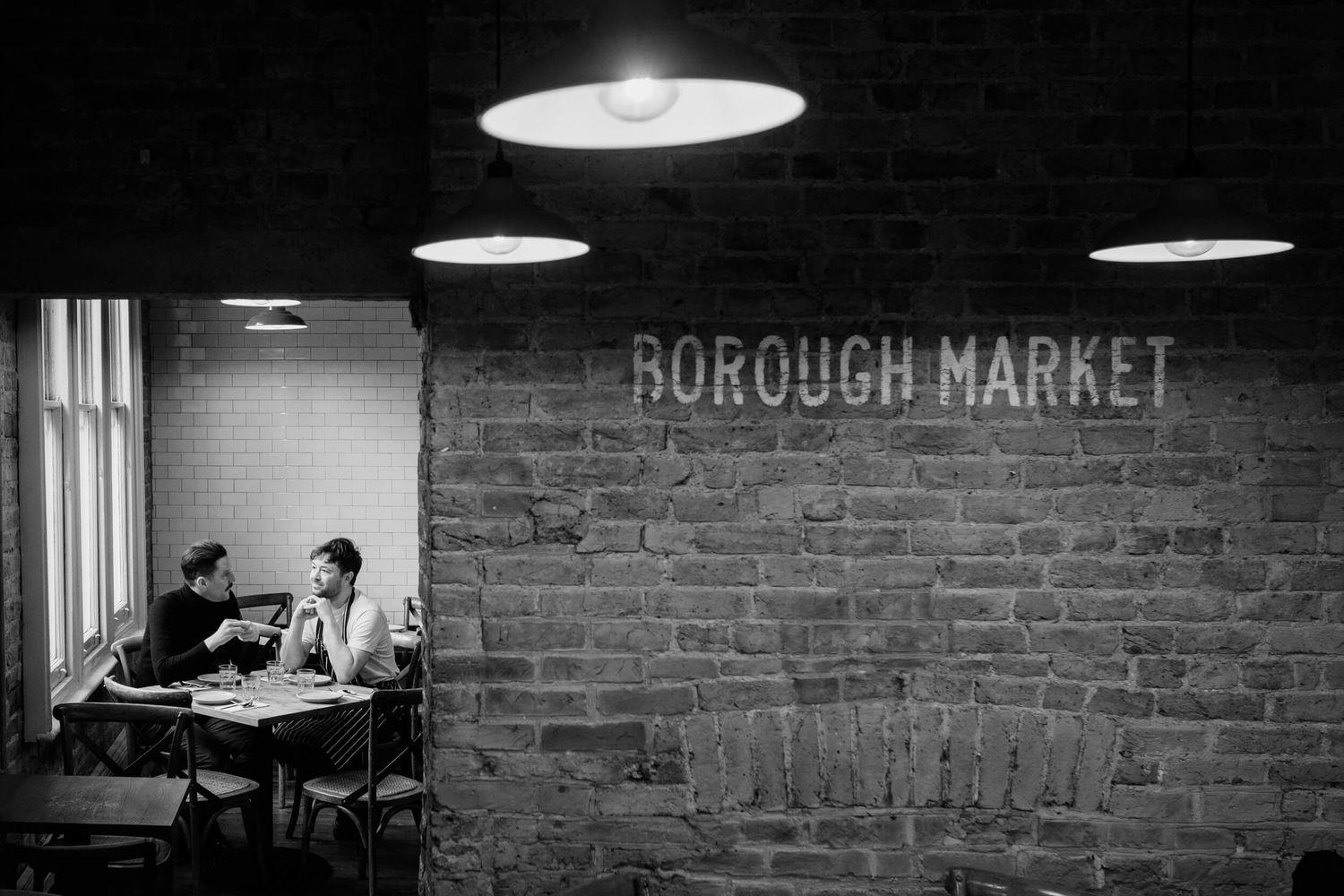Vintage black and white photograph showing people dining inside Borough Market with exposed brick walls and pendant lights.