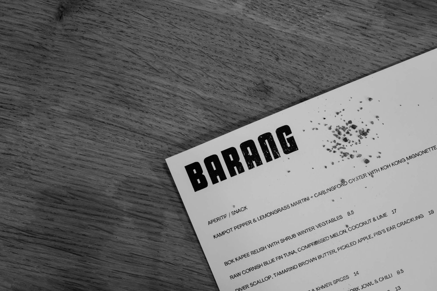 A black and white photograph shows a document with 'BARANG' text printed at the top on a wooden surface.