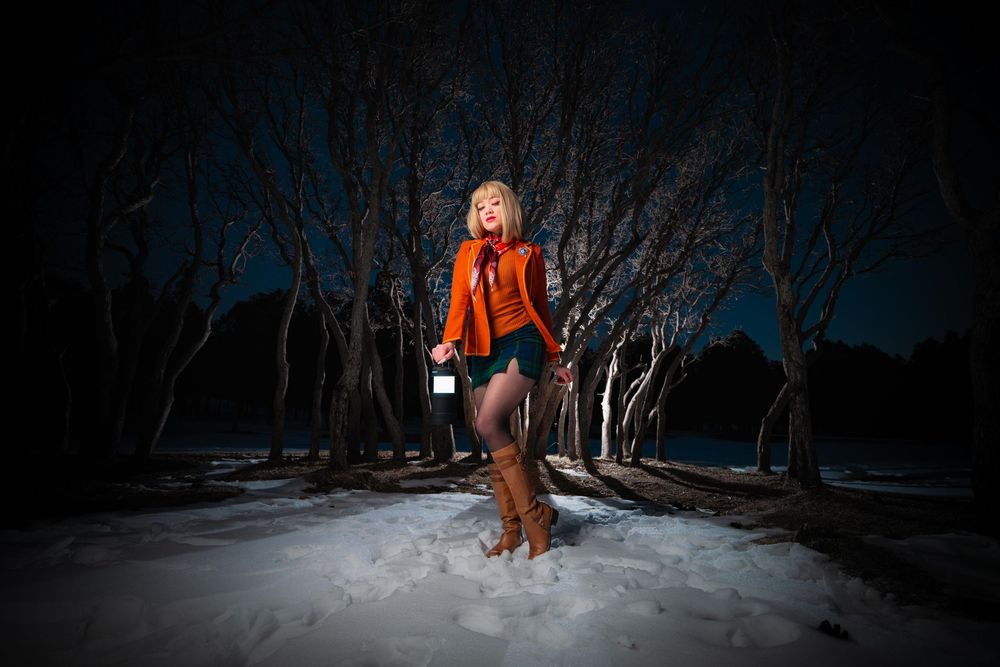 A person in an orange winter coat walks through a snowy forest landscape at night with dramatic lighting.
