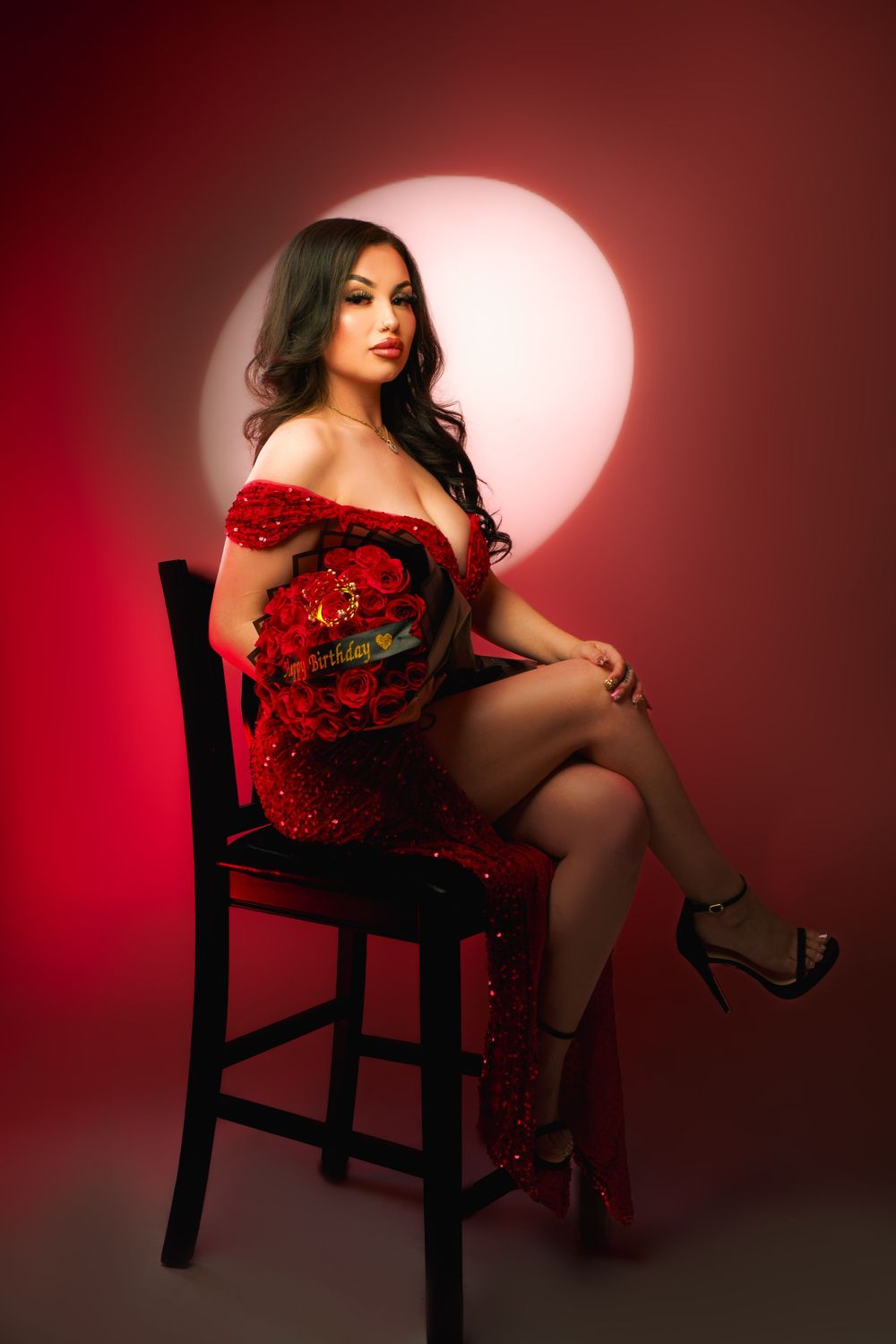 A glamorous portrait in red featuring a person in an off-shoulder dress seated on a dark chair against a dramatic circular light.