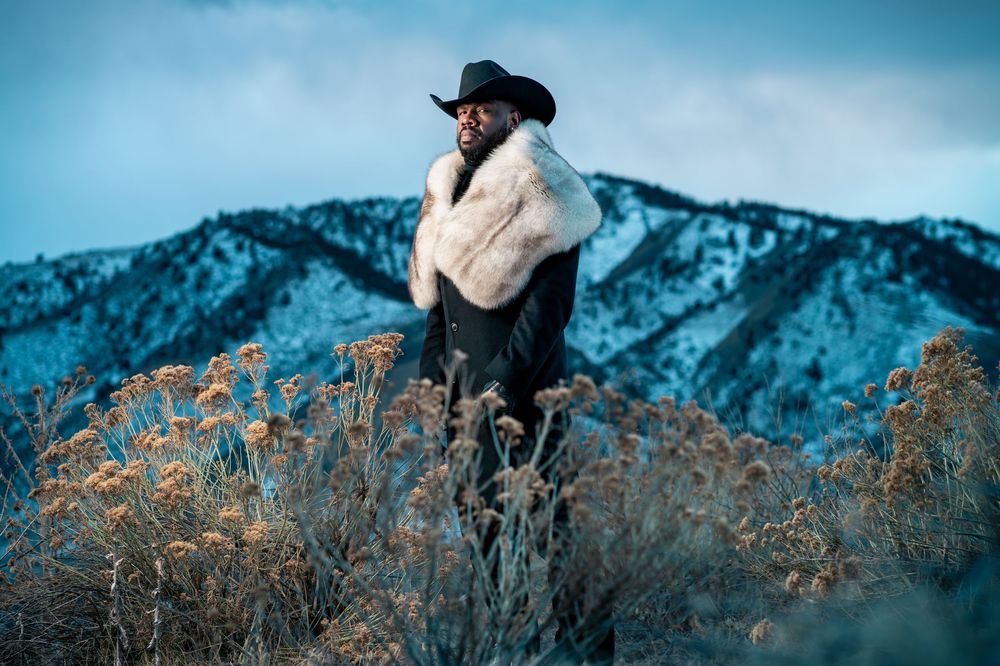 A person in a black hat sits on a rock overlooking snow-capped mountains during golden hour.