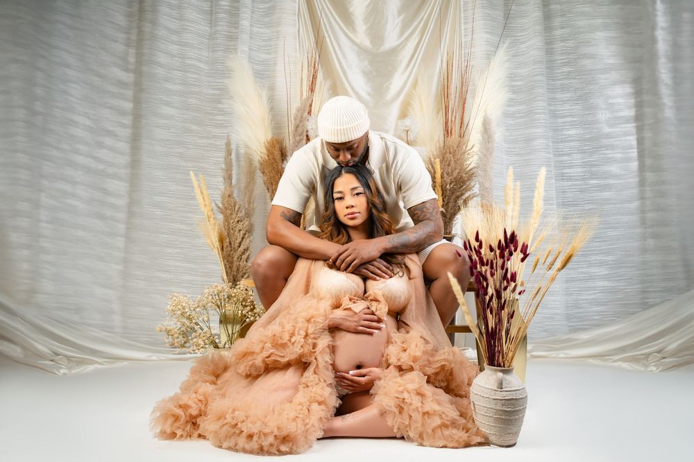 Romantic maternity photo with couple in white and peach tones surrounded by dried flowers and soft drapery.