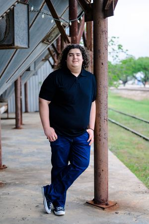A person in a navy shirt and blue pants poses against rustic metal columns on a covered porch walkway.