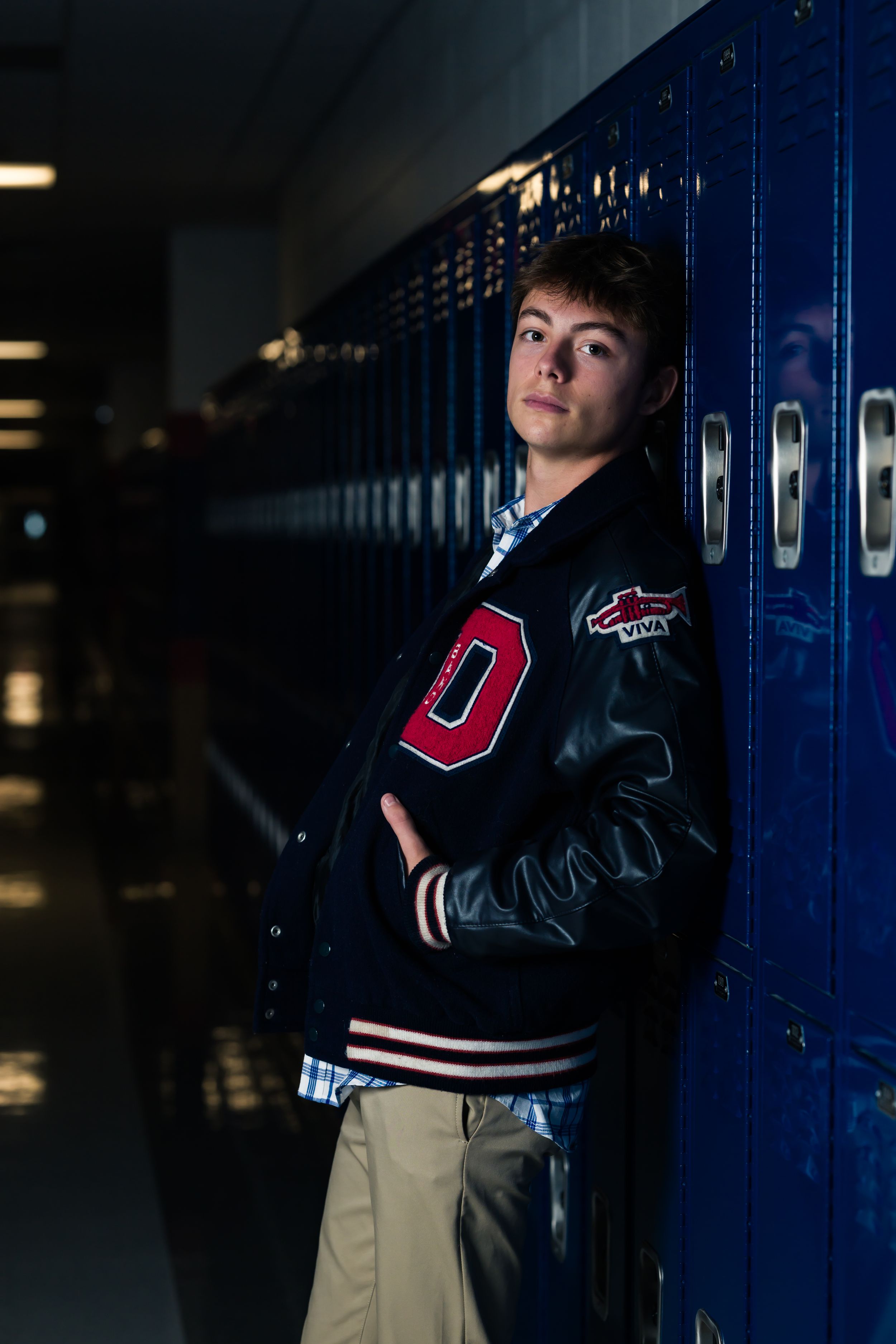 A student in a dark varsity jacket poses dramatically against blue school lockers in a dimly lit hallway.