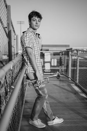 Black and white portrait of a student holding a trumpet while standing on a bridge walkway wearing plaid shirt and sneakers.