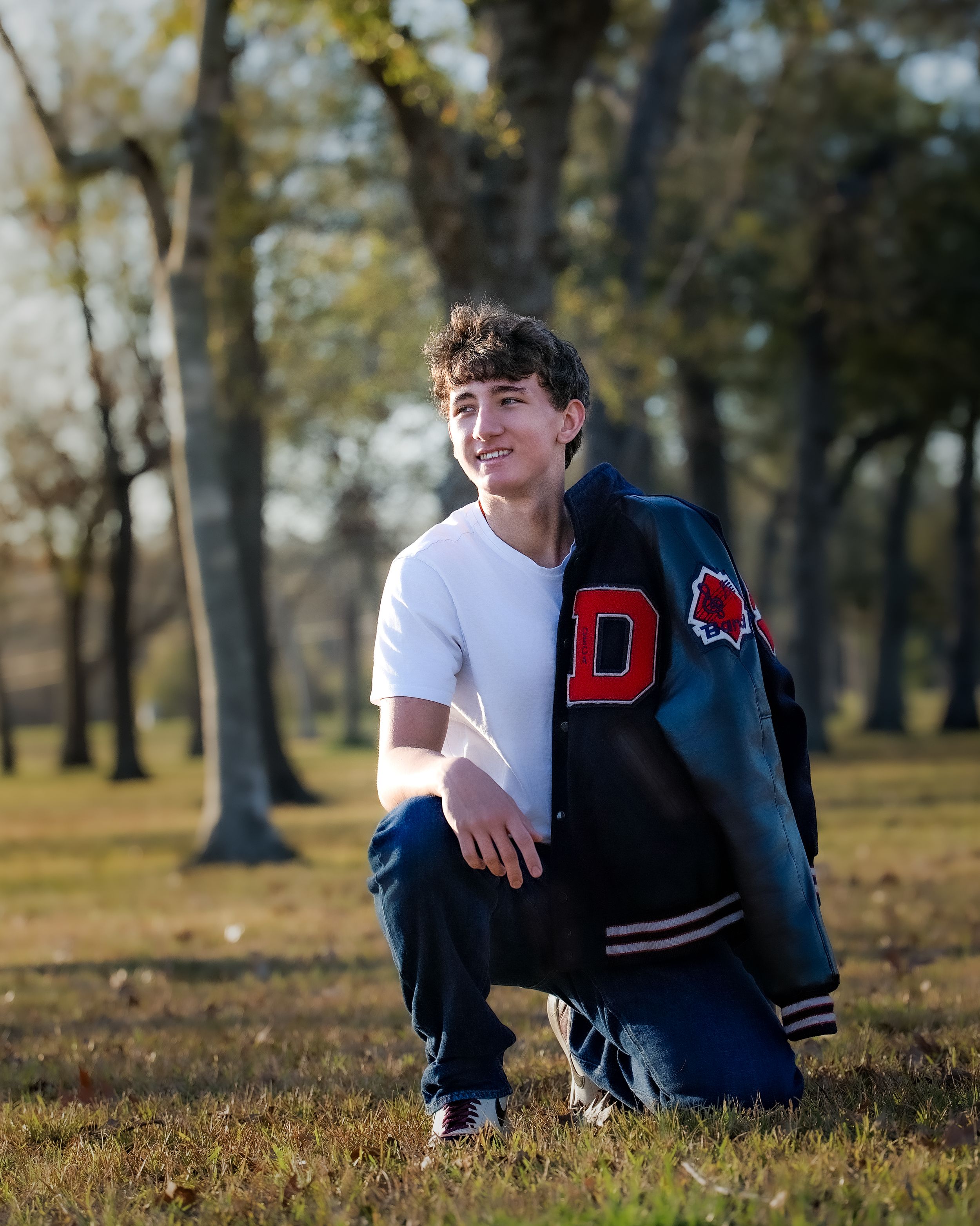 A young person in a varsity jacket sits on grass in a park setting during golden hour lighting.