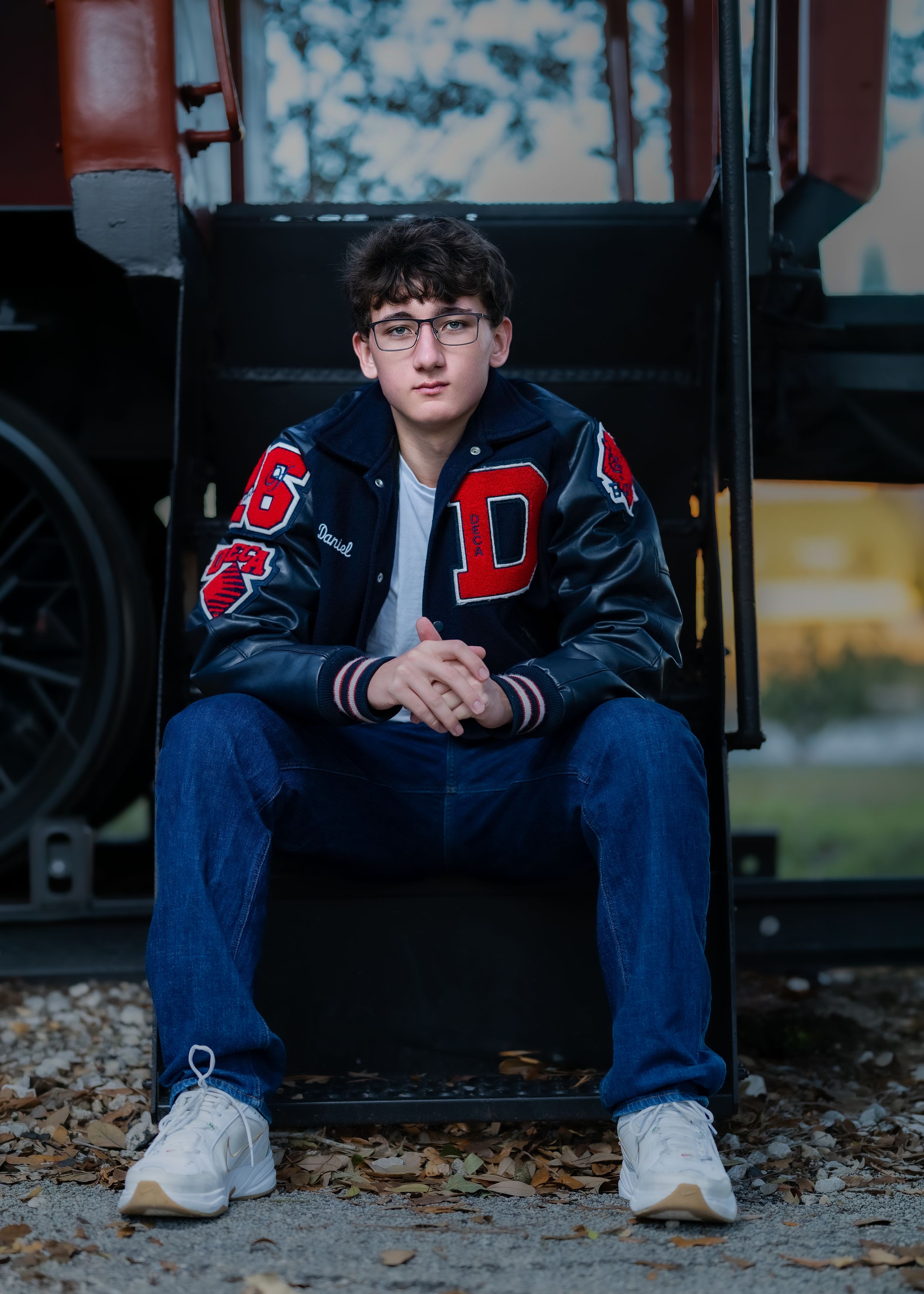 Student in varsity jacket sits on steps with autumn leaves scattered around in moody outdoor portrait setting.