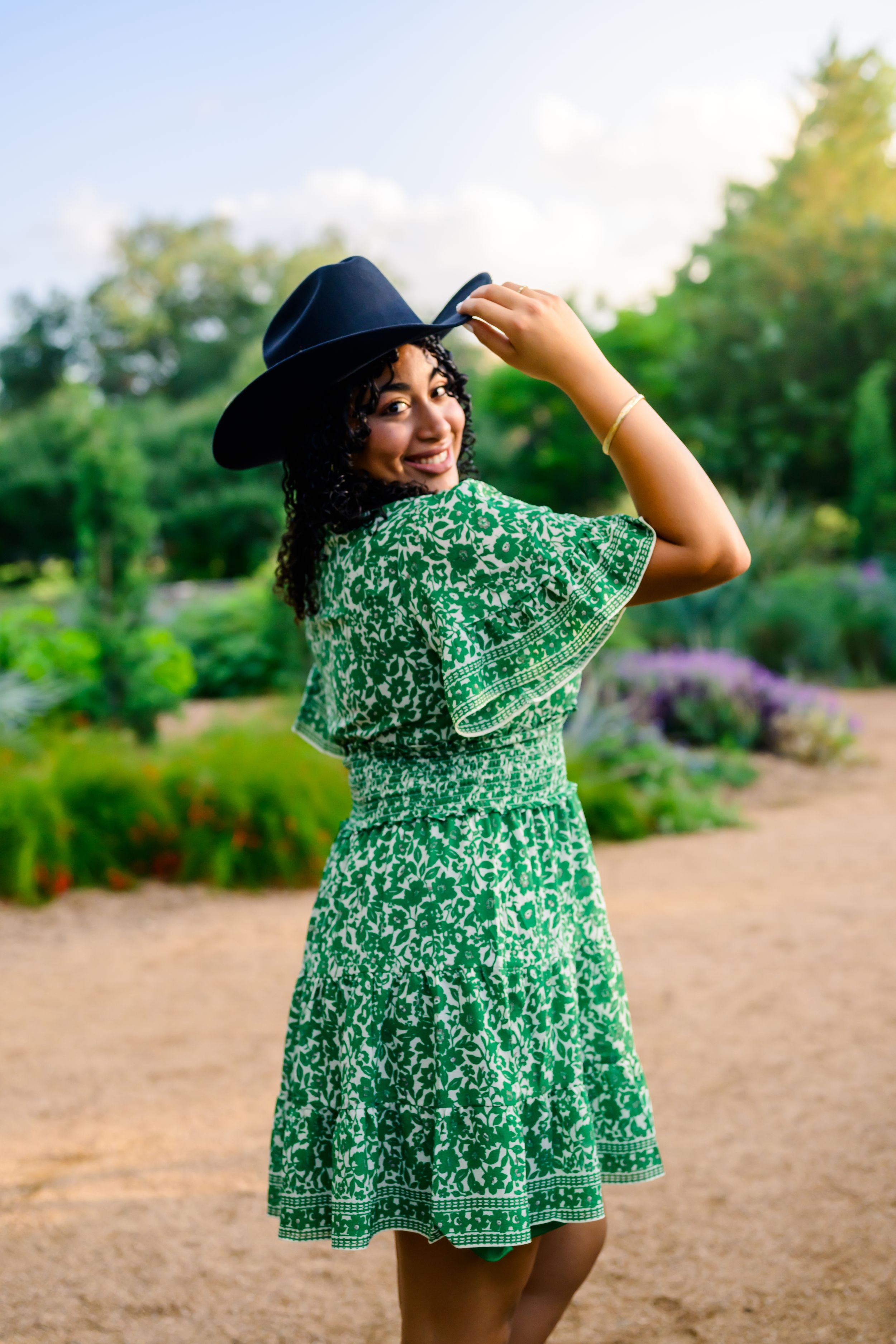 A person in a green floral dress and black hat poses on a garden path surrounded by summer blooms and greenery.
