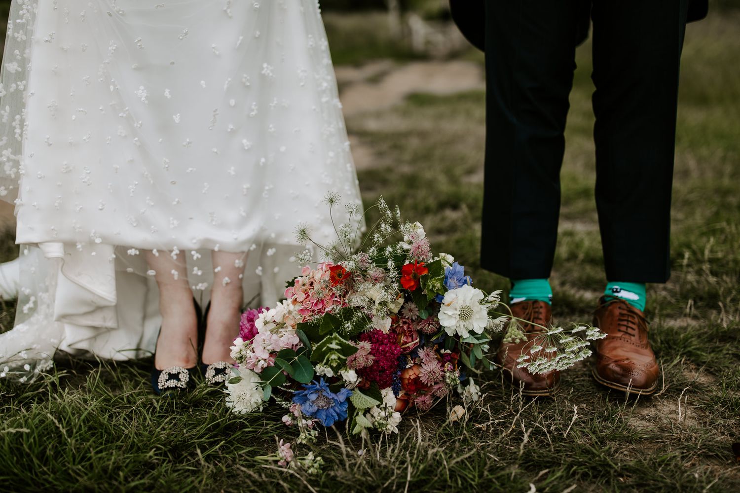A row of wedding guests' feet and shoes stand next to colorful floral arrangements on grass during an outdoor ceremony.