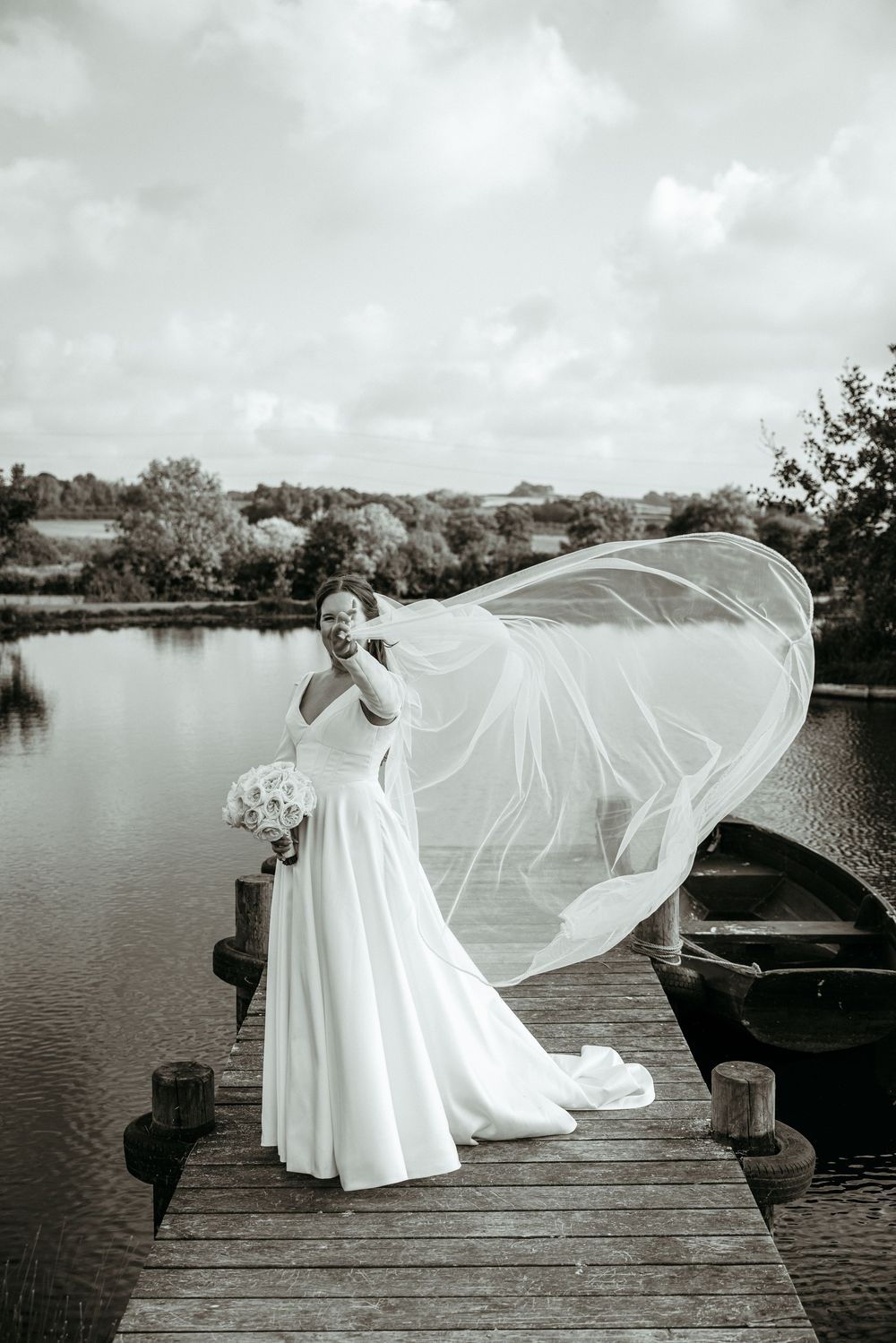 Black and white photo of flowing wedding dress and veil captured on lakeside dock.