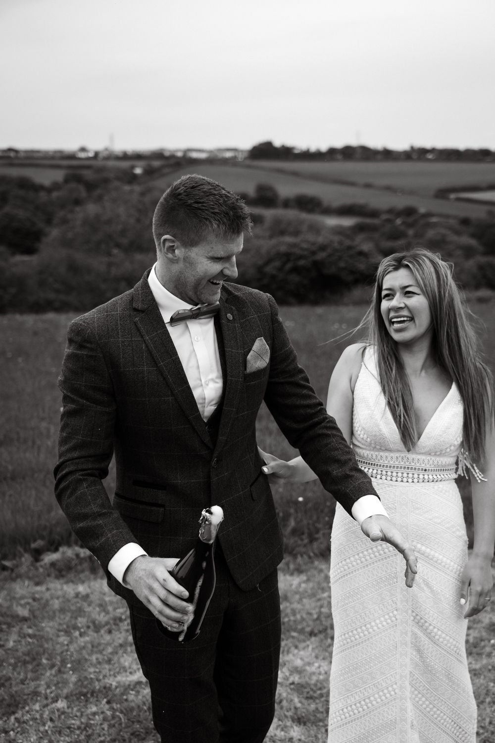 Candid moment of newlyweds laughing while holding champagne in a hilltop setting.