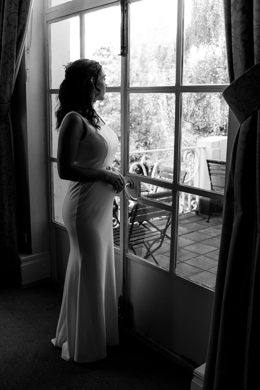 Black and white image of a bride silhouetted against large windows in a preparation room.