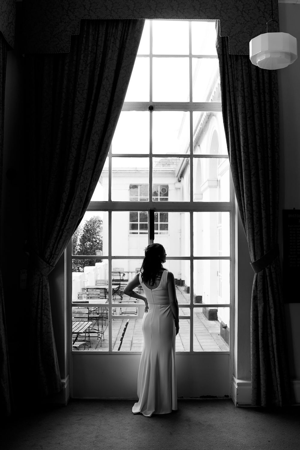 A silhouette stands by tall windows with dark curtains in a dramatic black and white composition.