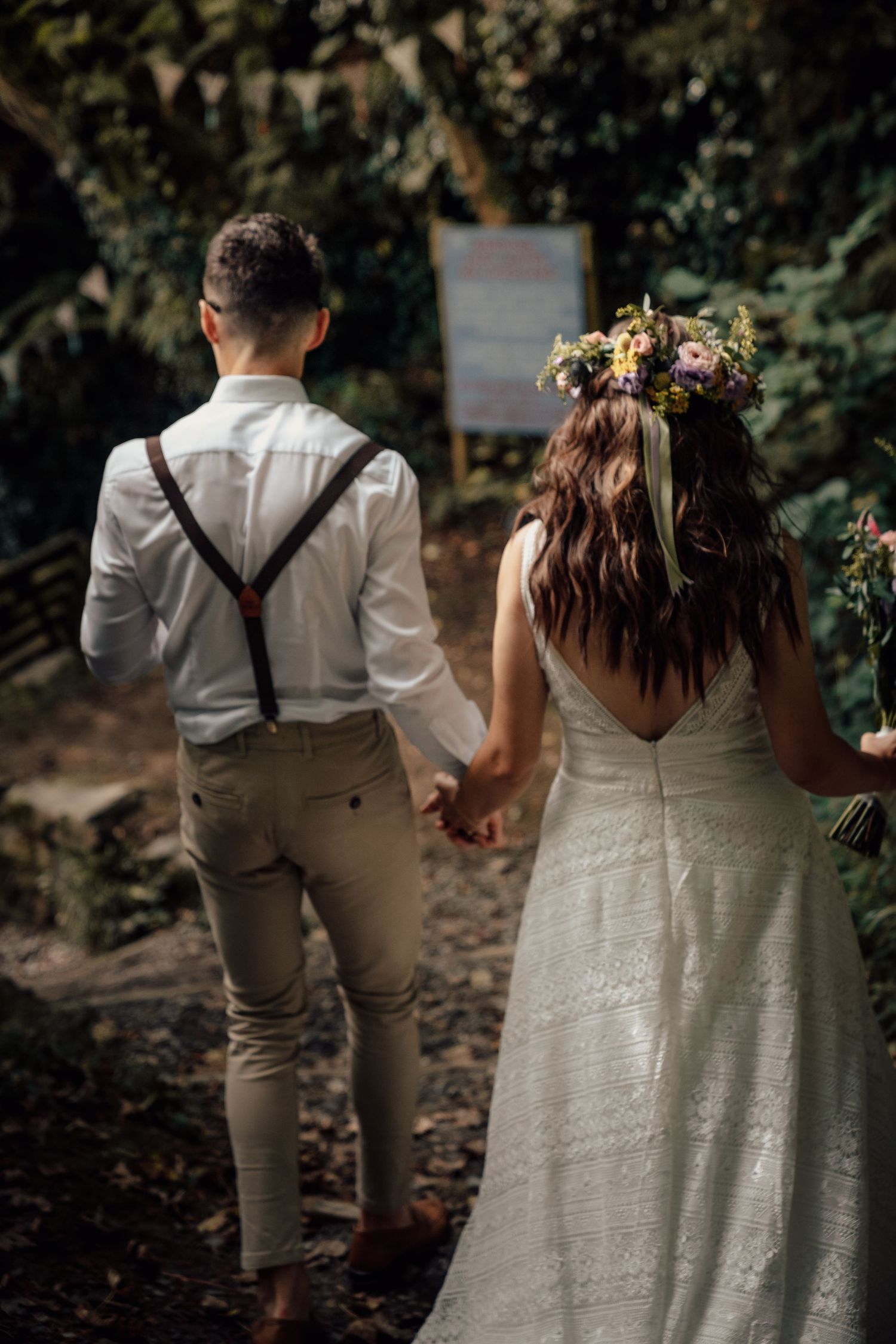 Couple in wedding attire walking hand in hand down a forest path.
