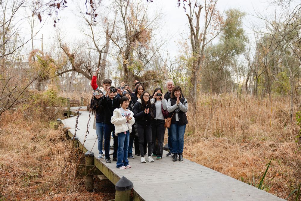 A group stands together on a wooden boardwalk path through autumn wetlands and tall grass on a cloudy day.