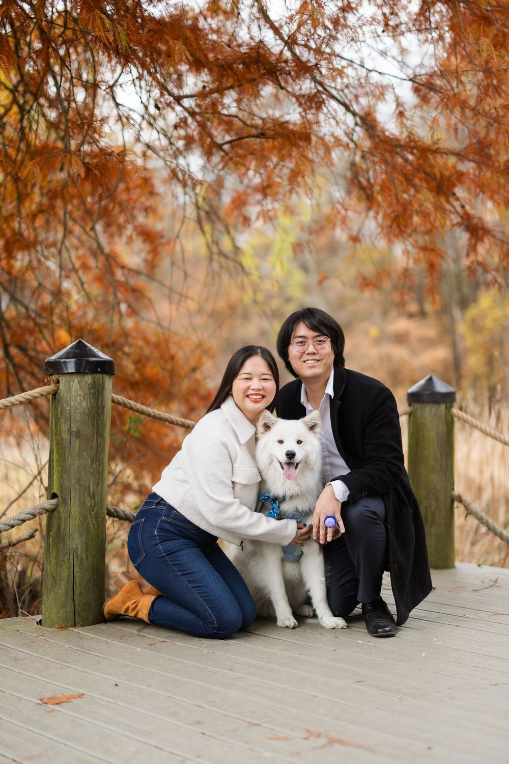 A white Samoyed dog poses with two people on a wooden bridge surrounded by autumn foliage.