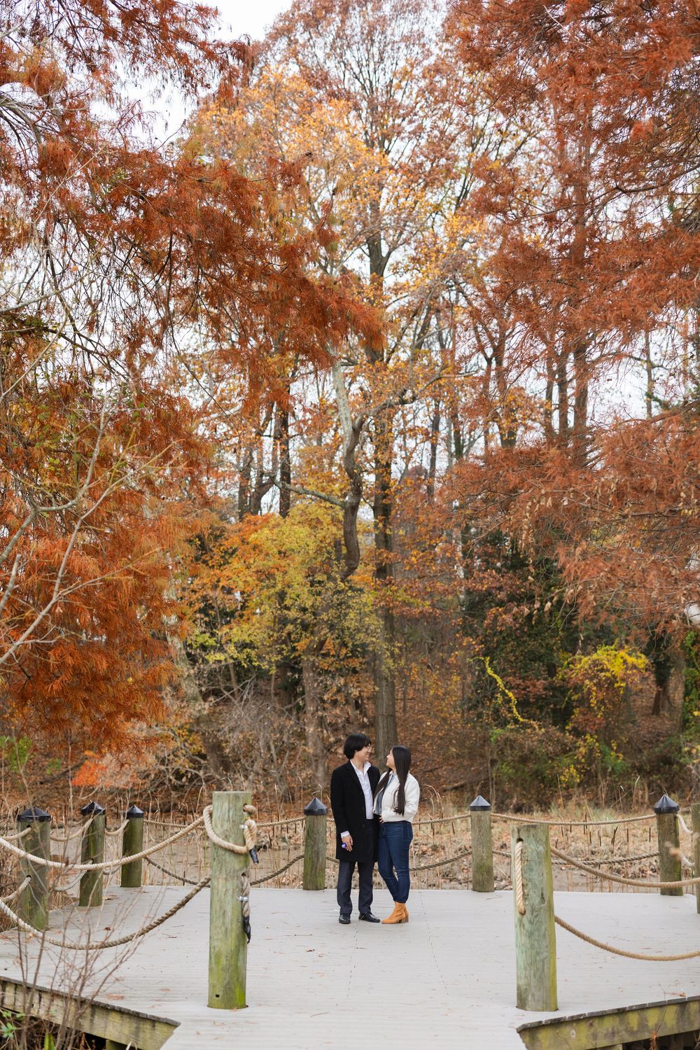 A couple walks along a snowy path surrounded by autumn trees and wooden posts.