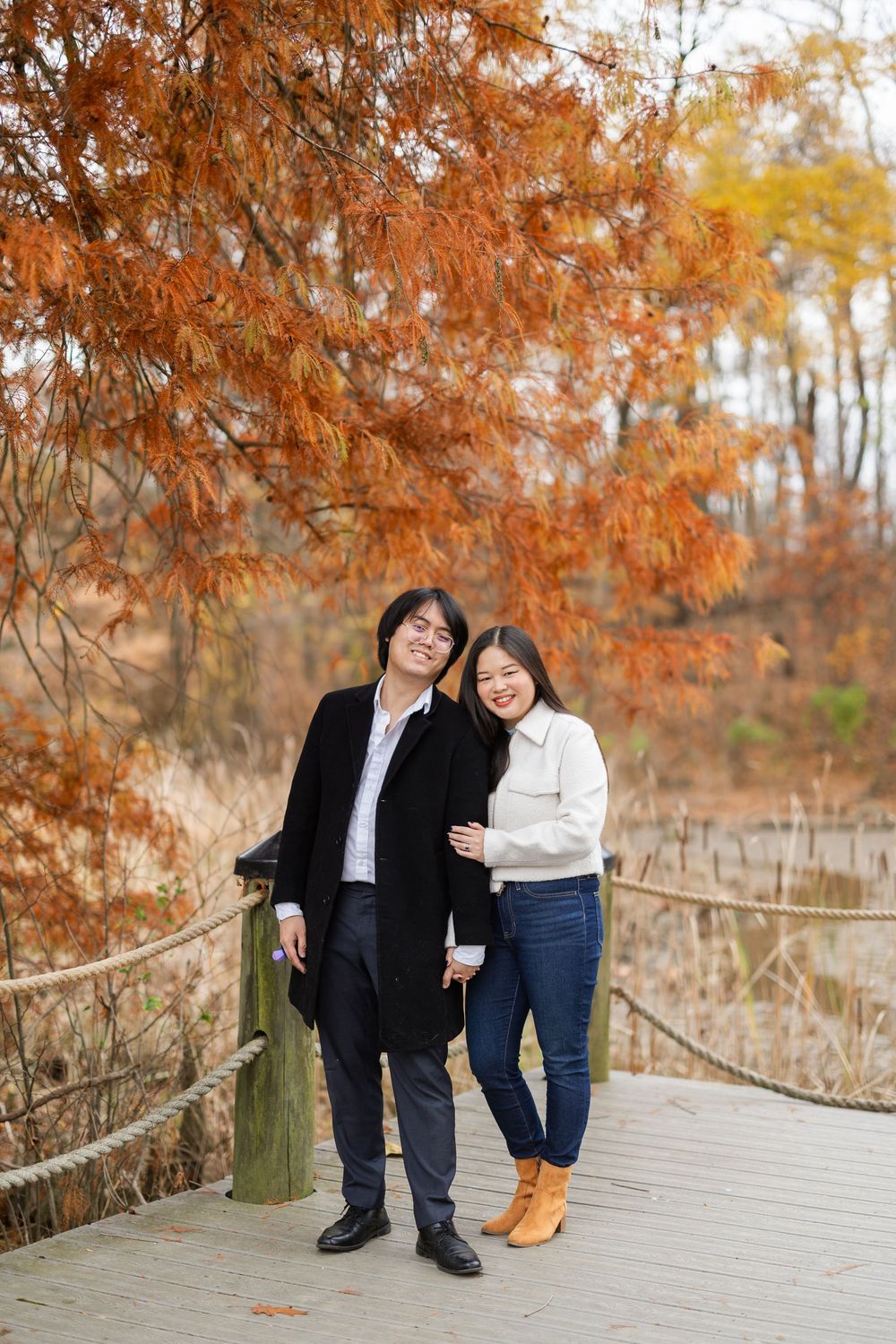 A couple walking and posing along a wooden path with orange fall leaves overhead.