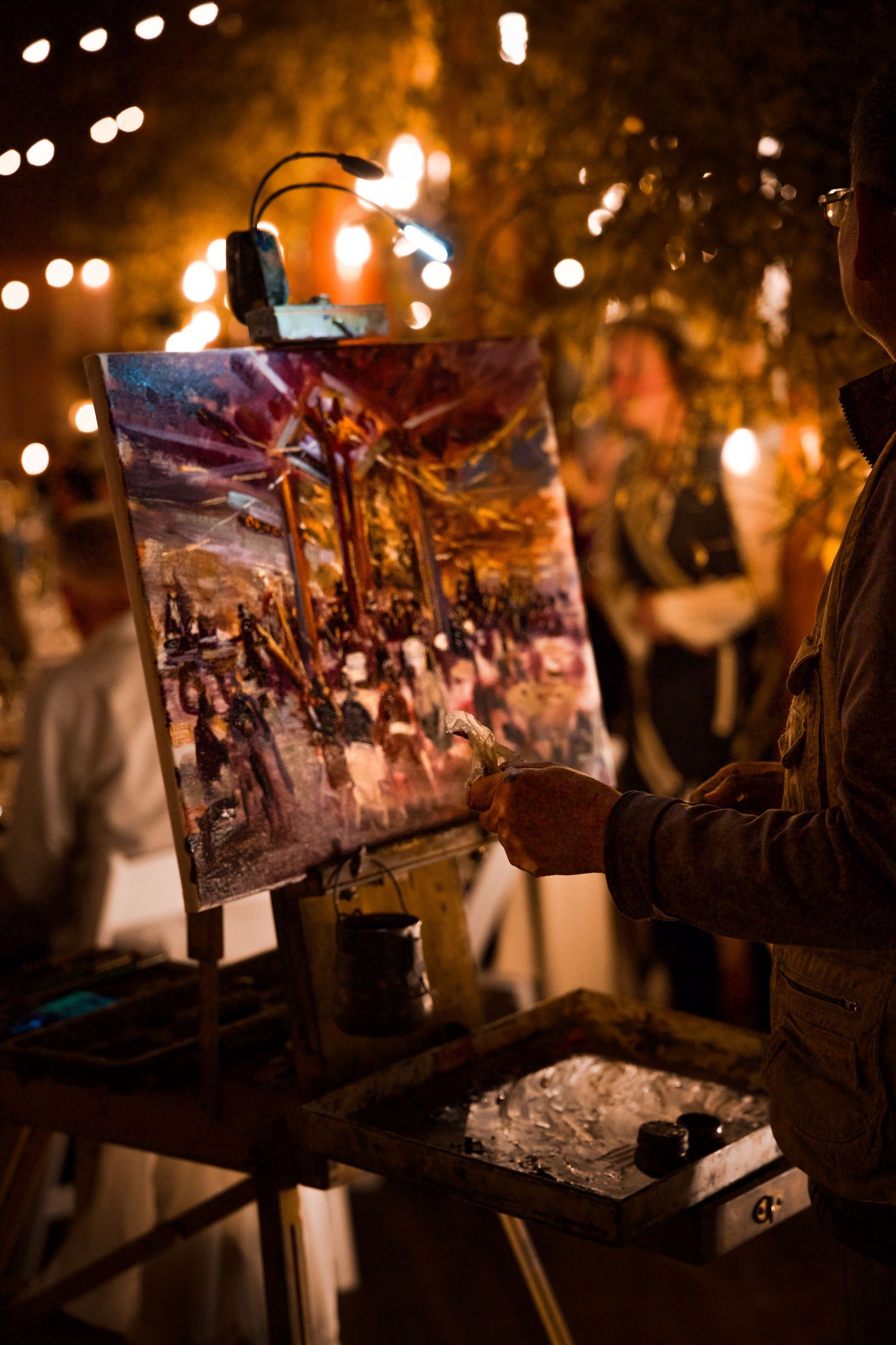 Artist painting at an outdoor evening event with string lights and an illuminated canvas on display.