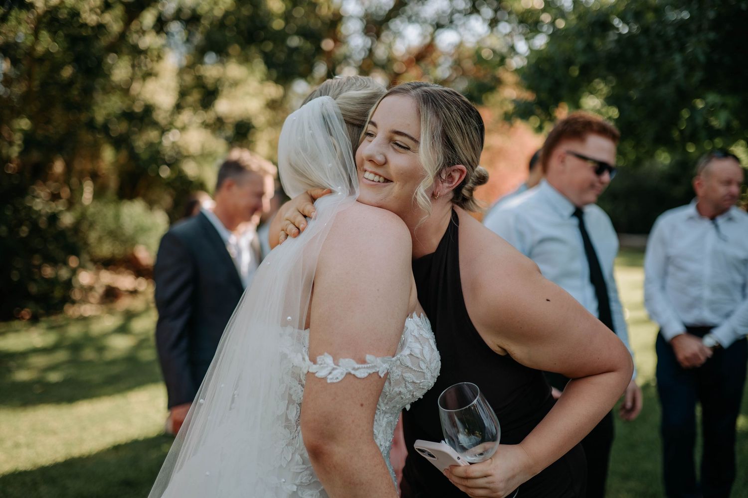 Wedding guests gather outdoors for a celebration in a sunlit garden setting.
