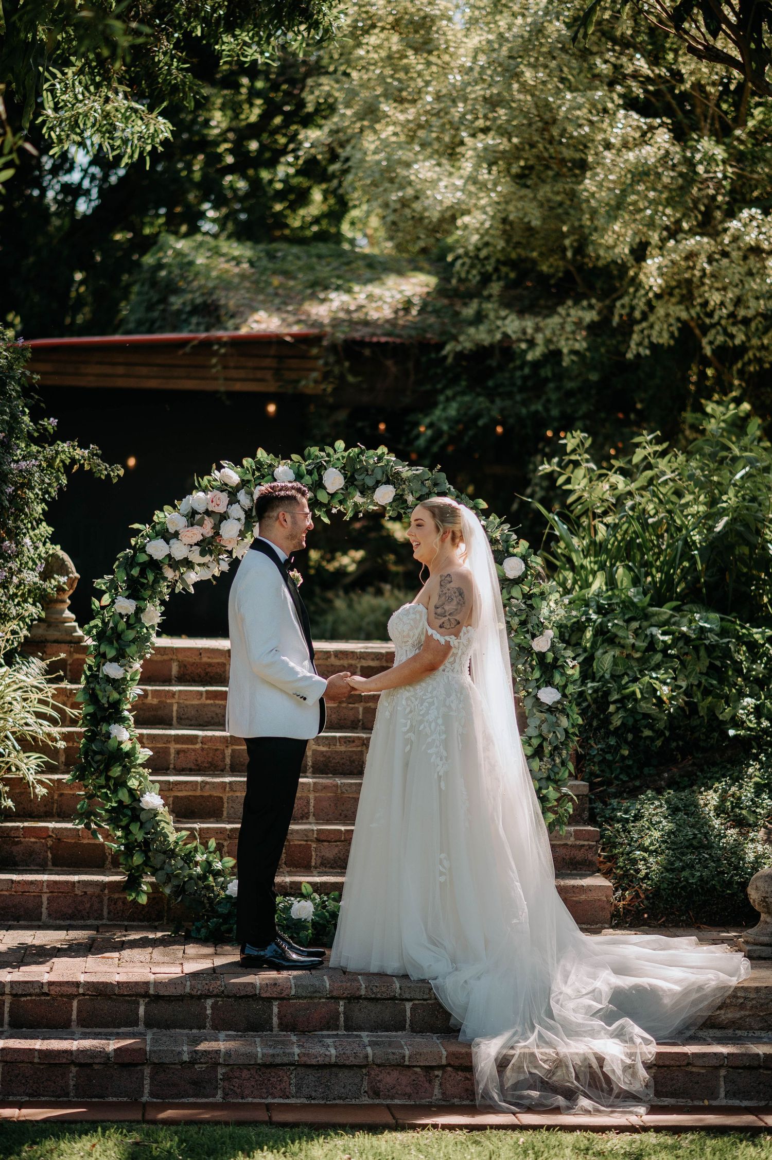 A romantic wedding first look moment unfolds on rustic brick steps surrounded by lush greenery and a floral arch.