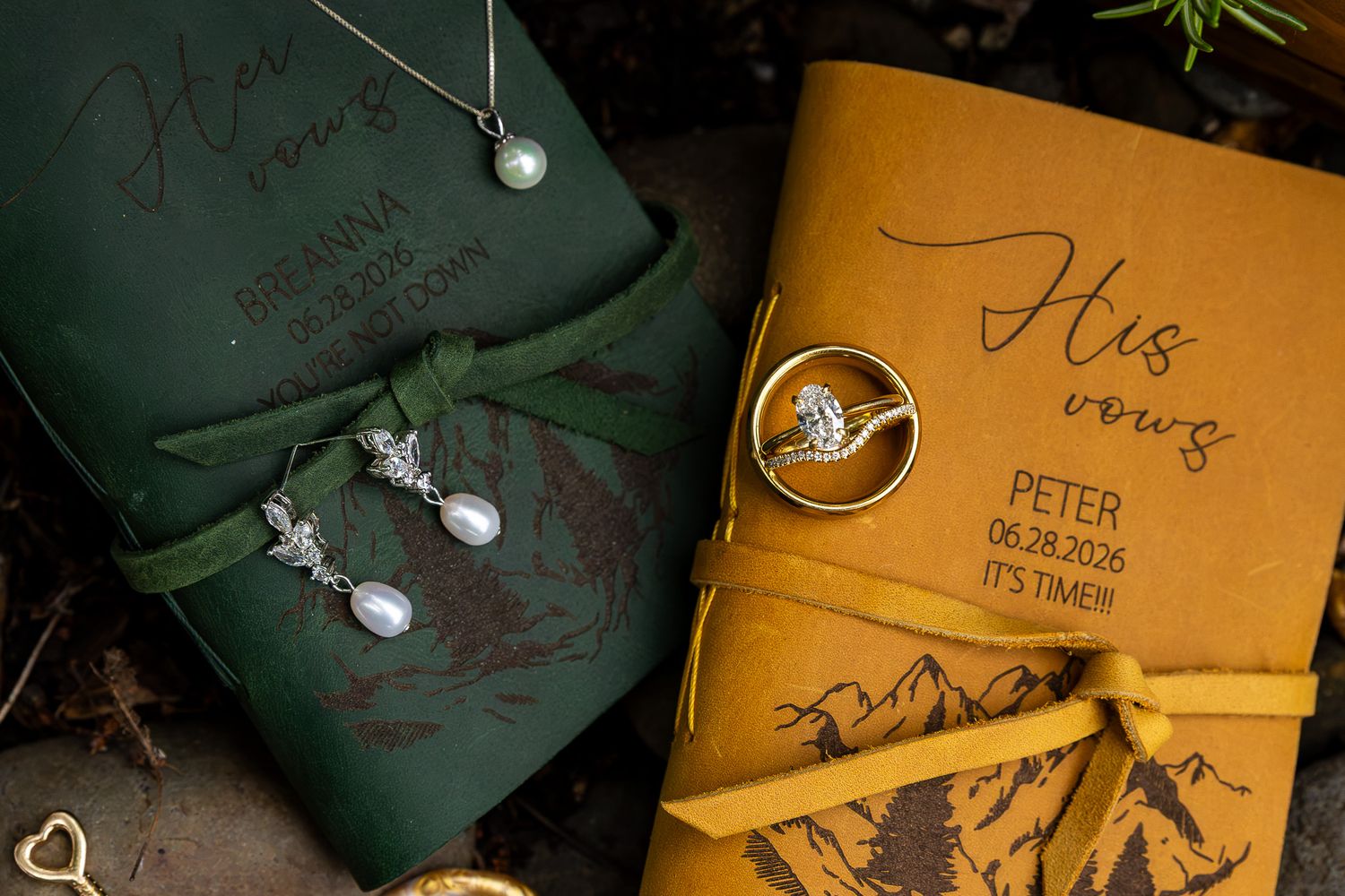 Green and mustard yellow leather vow books with engraved names, pearl earrings, and diamond ring on rustic background.