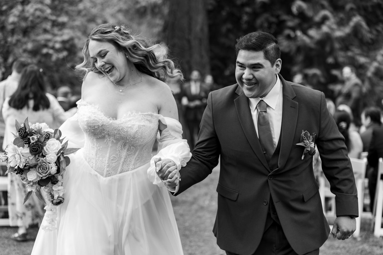 Black and white photo of a joyful bride and groom holding hands, walking down the aisle after their outdoor wedding ceremony.