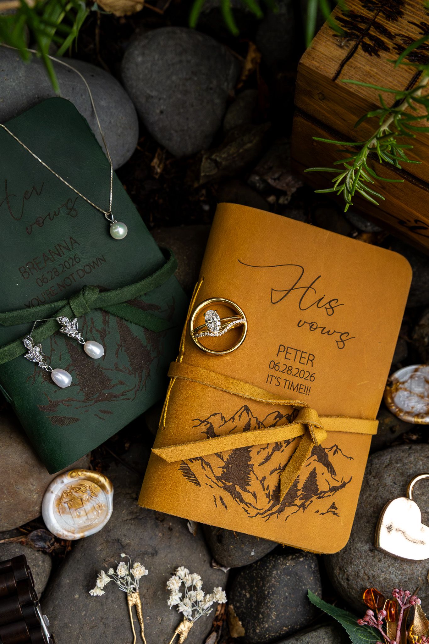Personalized leather vow books in green and mustard yellow with mountain engravings, wedding rings, pearl earrings, and necklace on rocks.