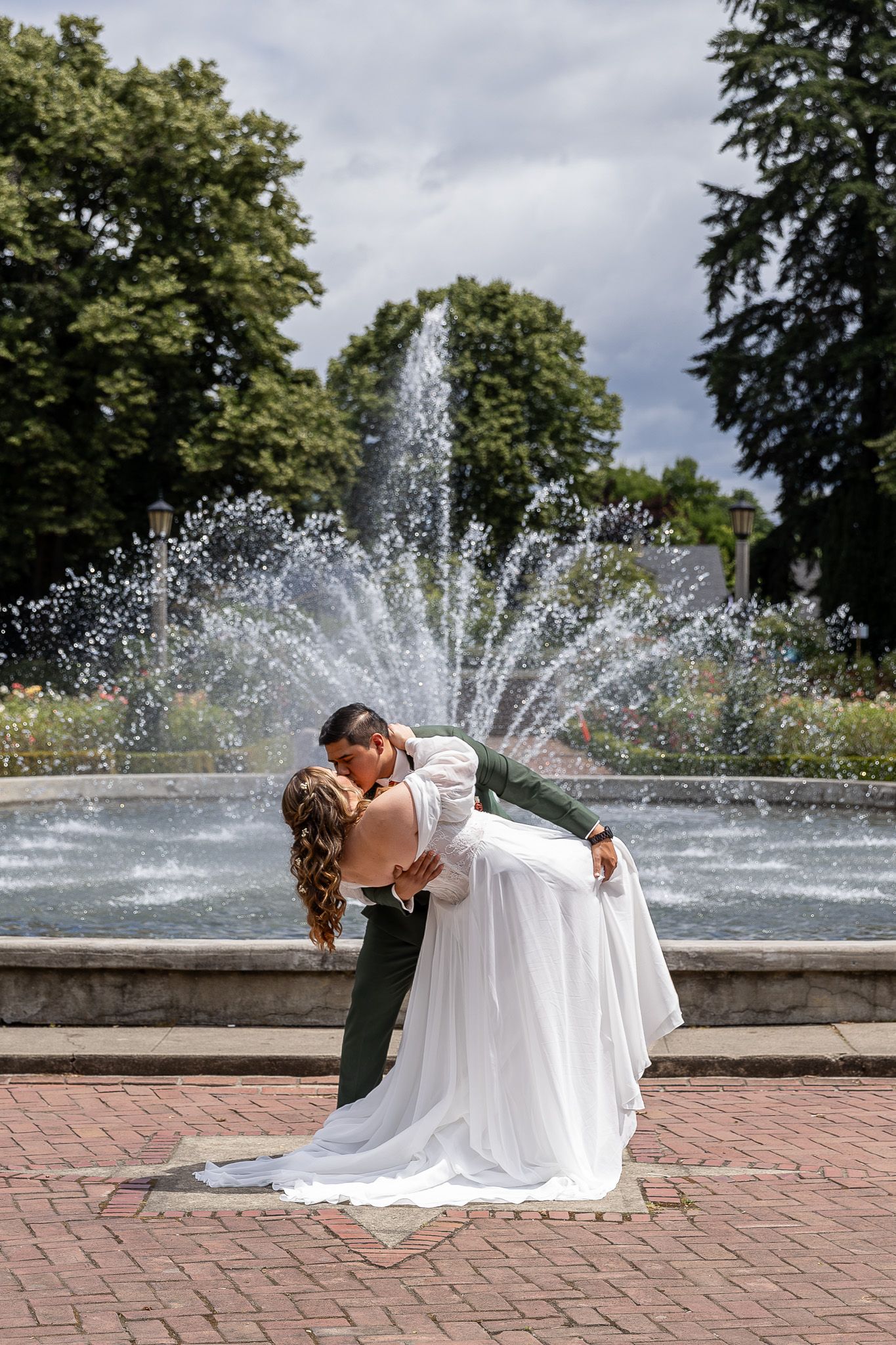 Bride and groom share a romantic dip kiss in front of a splashing fountain in a lush green park on their wedding day.