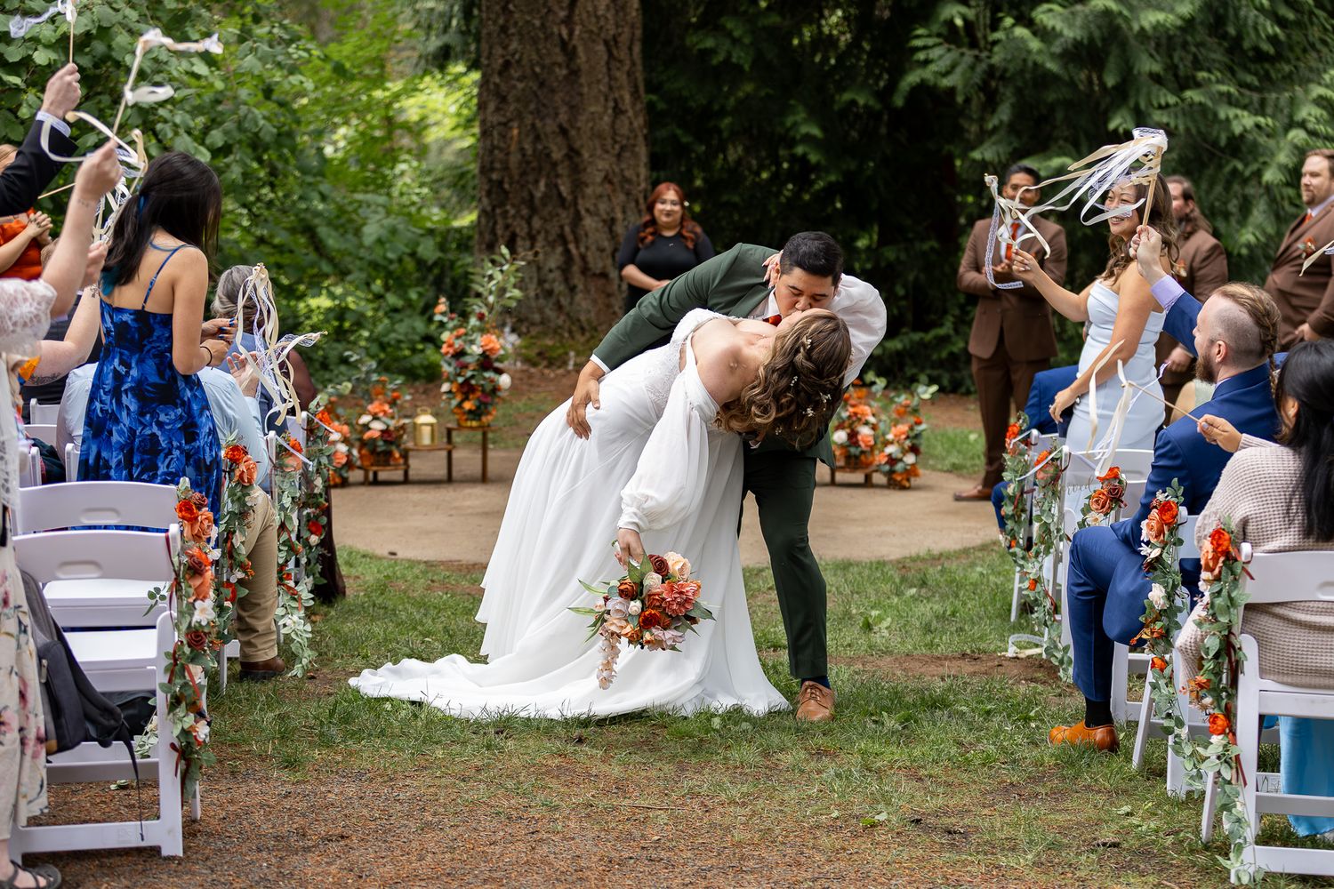 Couple shares romantic dip kiss at outdoor wedding ceremony aisle surrounded by guests waving ribbon wands.