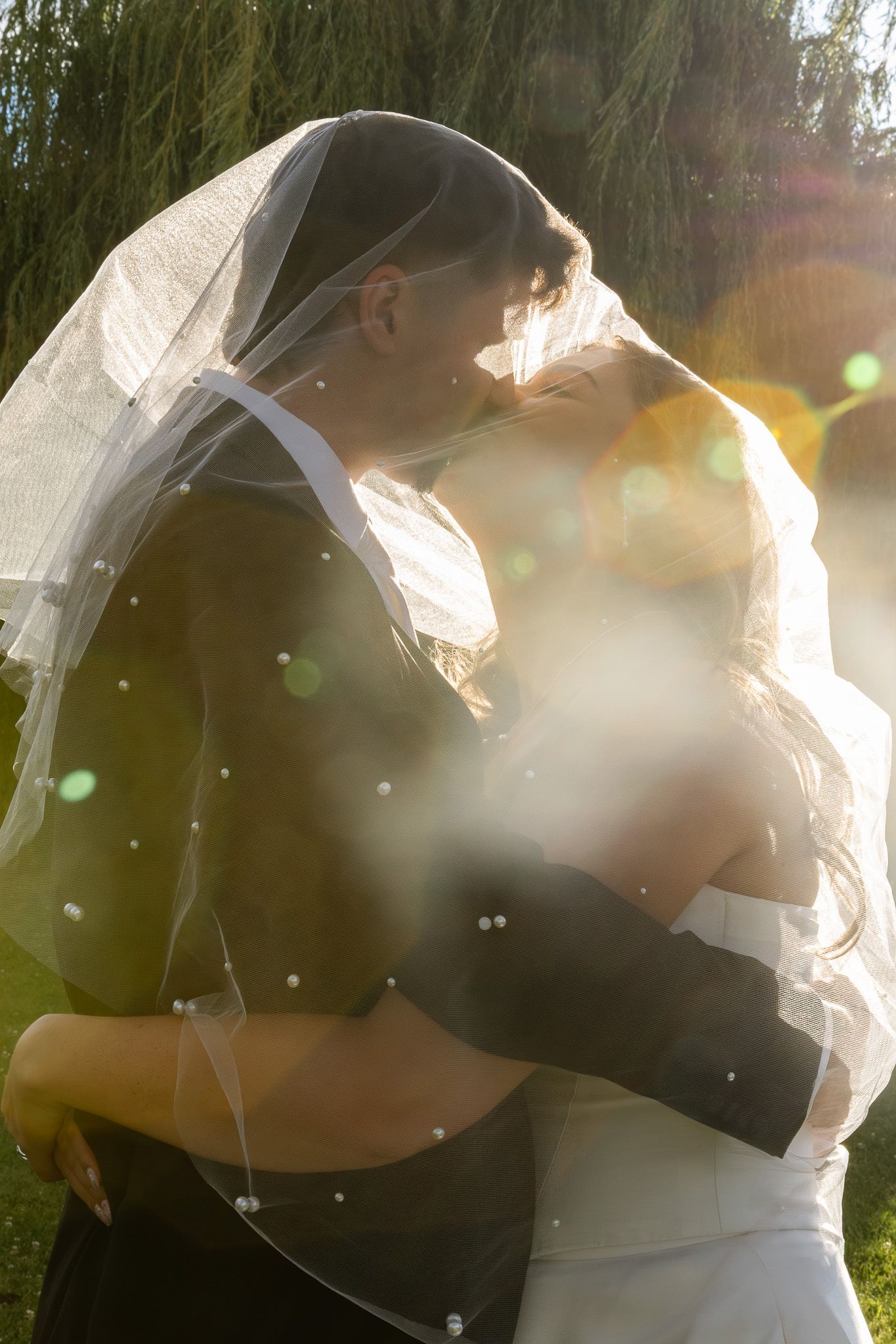 Bride and groom share a romantic kiss under a veil with golden sunlight and bokeh effects outdoors.