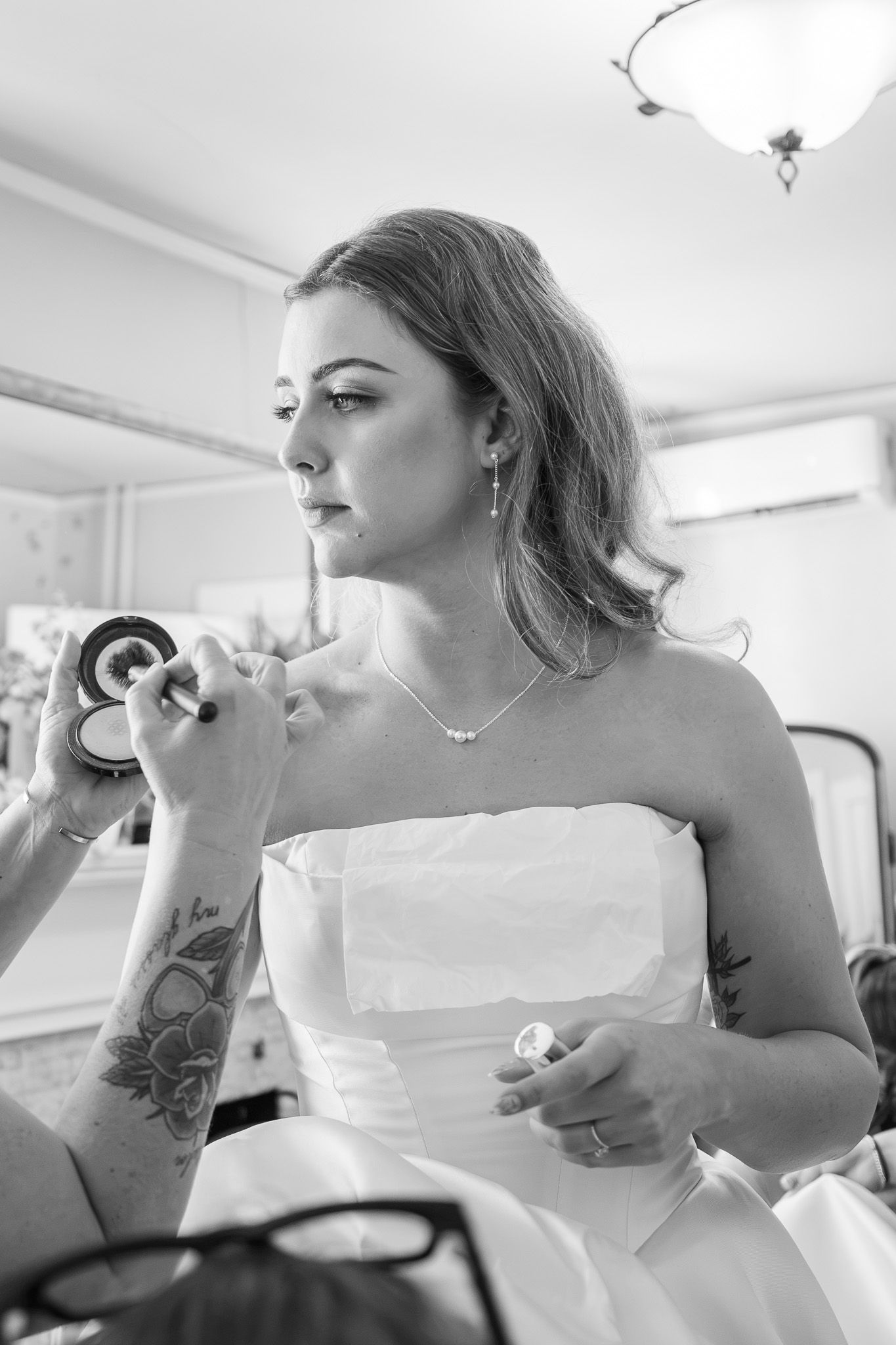Black and white photo of a bride in a strapless gown getting makeup applied by a tattooed artist before her wedding.