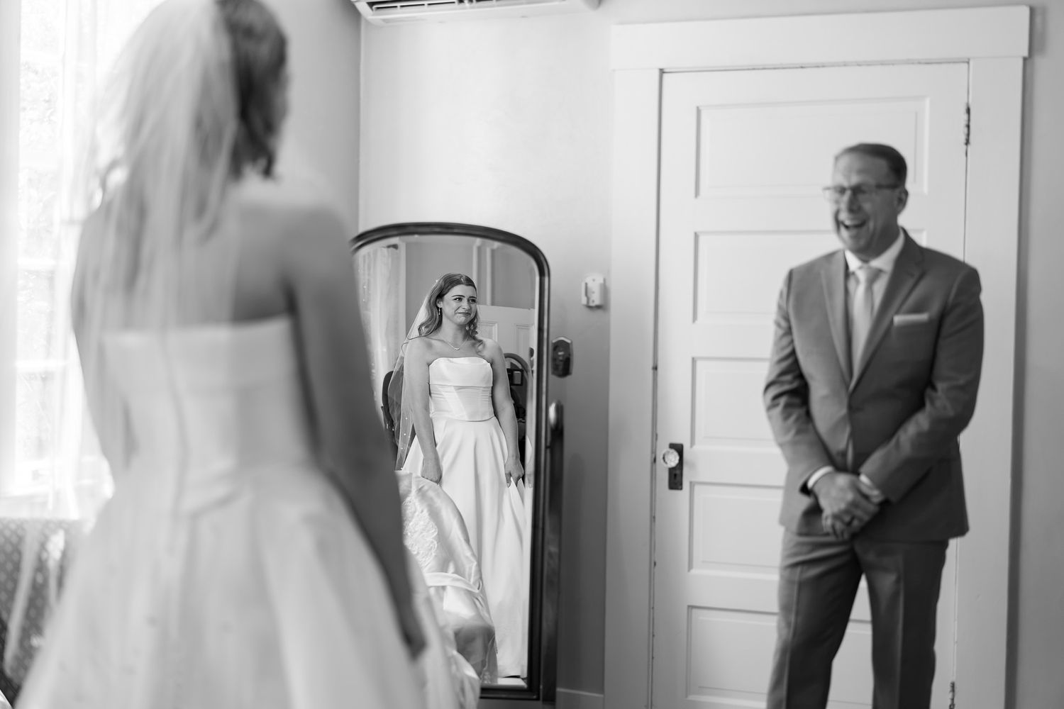 Black and white photo of a bride in a wedding dress seeing her reflection in a mirror while a man in a suit looks on.
