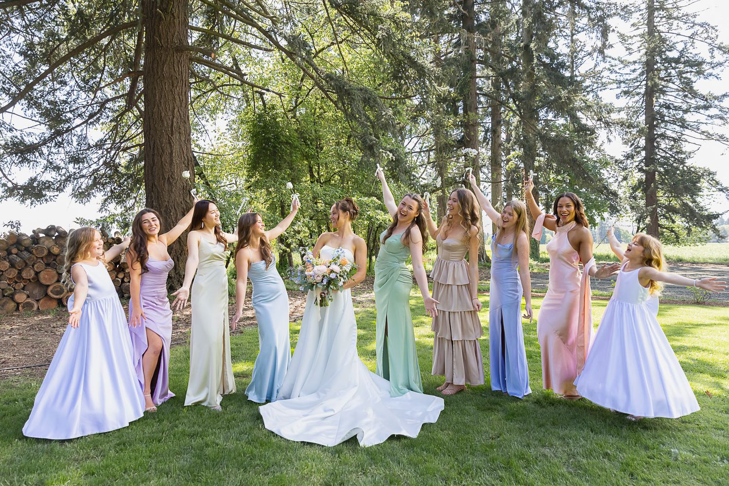 Bride in white gown poses with bridesmaids in pastel dresses outdoors under tall trees on a sunny day.