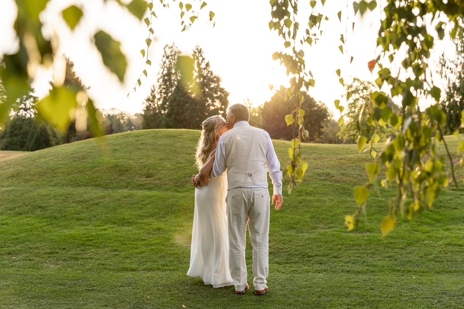 Couple in white wedding attire walking hand-in-hand on lush green lawn at golden sunset.
