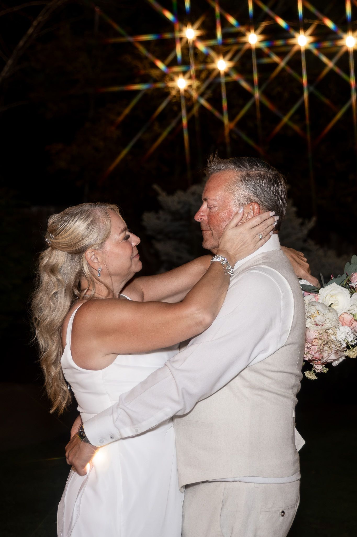A couple shares a romantic first dance at their wedding reception under sparkling string lights at night.