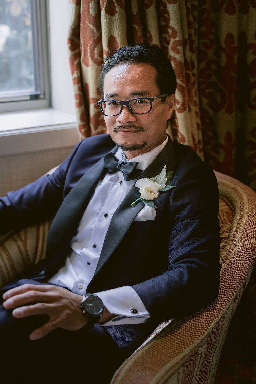 A person in a black tuxedo and bow tie sitting in an upholstered chair.