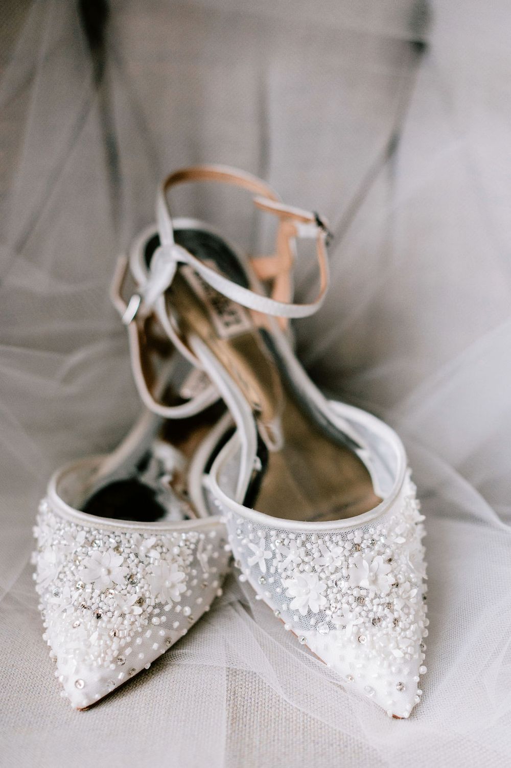 Delicate white bridal shoes adorned with crystal embellishments and beading displayed on tulle fabric.