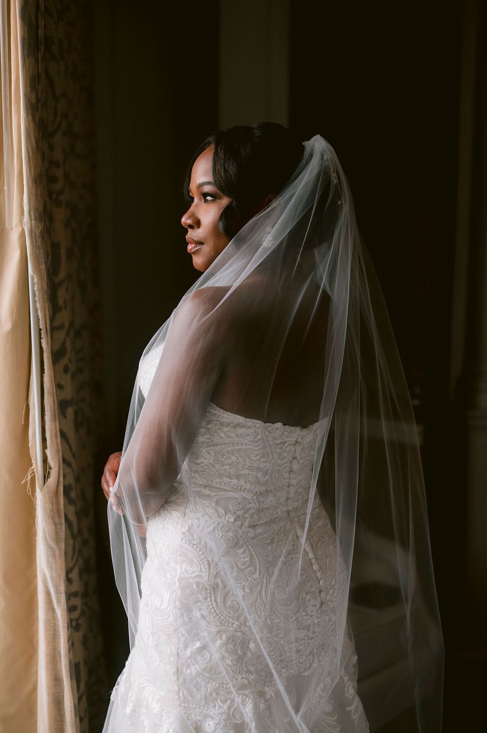 Elegant wedding dress with delicate lace details and flowing veil captured in dramatic window lighting.