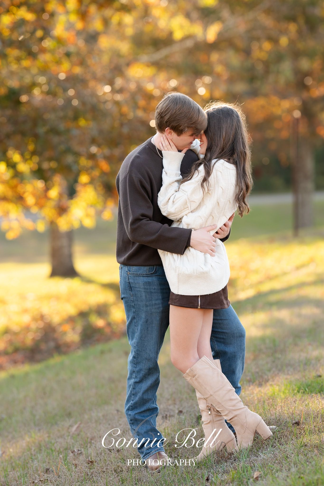 Romantic couple embraces in autumn setting with golden sunlight filtering through fall foliage in background.