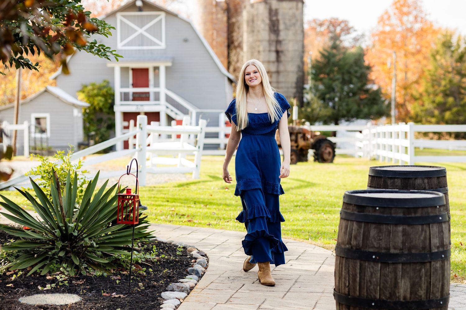 A person in a navy blue dress poses near rustic wooden barrels and autumn foliage in a garden setting.